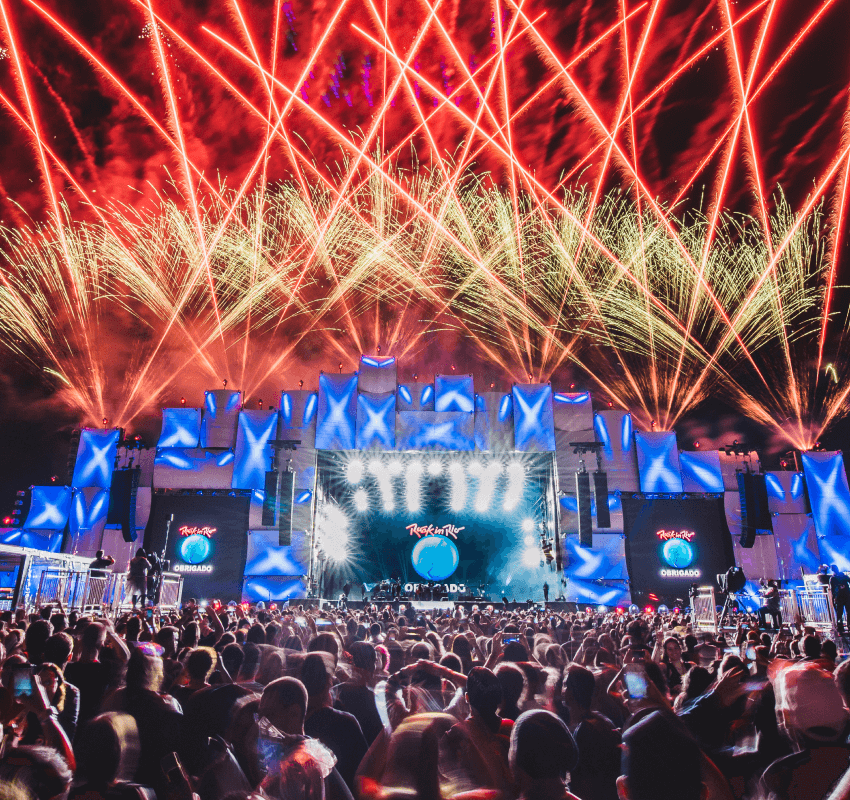 Festival de música Rock In Rio sendo finalizado. Fogos de artifício explodem acima do palco, em frente à plateia. No telão, logotipo do Rock in Rio e abaixo lê-se: obrigado.